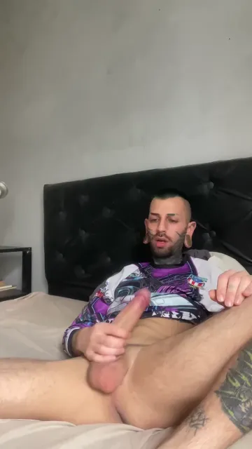 Tattooed Man Masturbates on Bed with Legs Apart